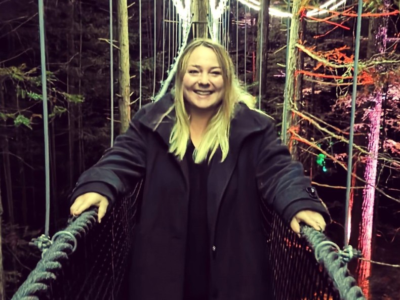 Jemma has blonde shoulder length hair. She is wearing a black coat and is standing on a bridge. 