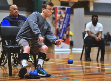 Young man with mobility impairment is sitting on a chair rolling his boccia balls.