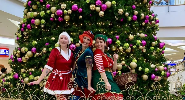 Three of santas elves are sitting beneath a large christmas tree