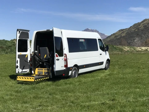 Mobility van with dropping ramp out the back is pictured in a feild with mountains behind