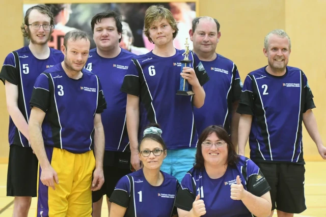 A team from the Fulton Hogan basketball league poses with a trophy. They are wearing purple and black t-shirts.