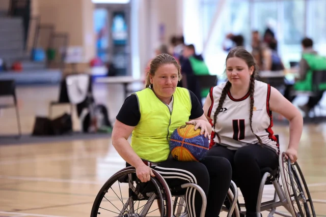 A female wheelchair basketball player in a bright yellow vest holds the ball while wheeling around a player in a white singlet.