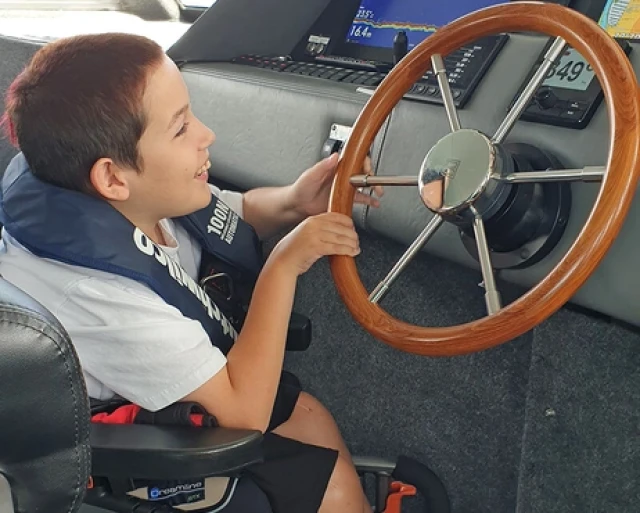 Kid in a powerchair and lifejacket at wheel of the boat