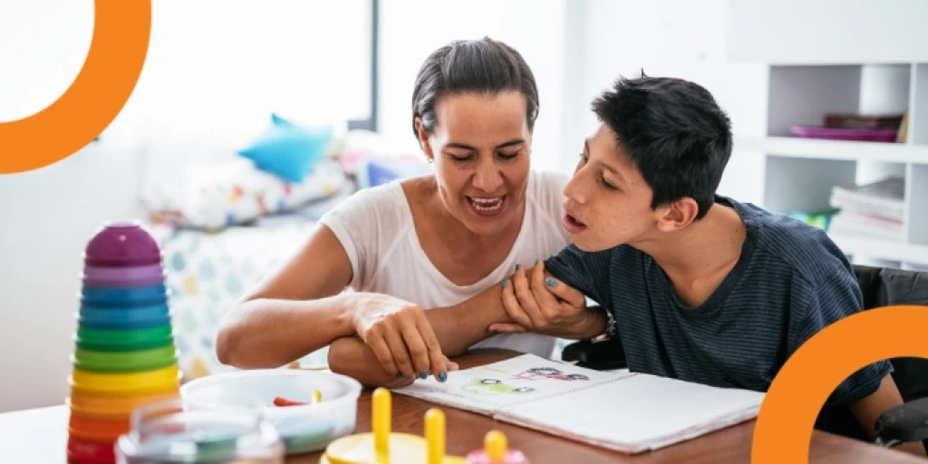A disabled boy sits at a table with his mother, looking at a book