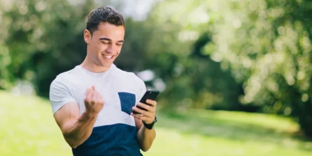 Young man in a blue and white shirt looking at his phone, pumping his fist.