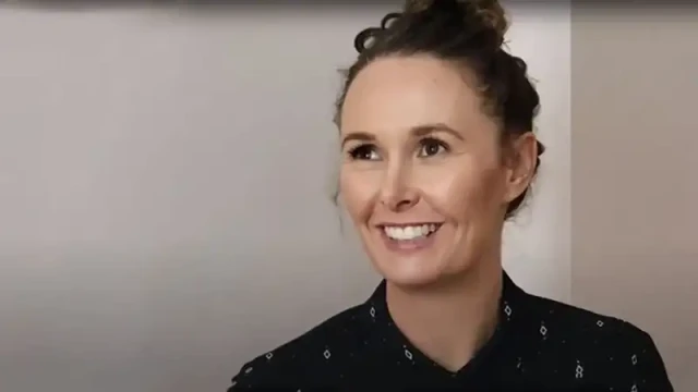 Dr Dr Carey-Ann is on the right side of 40 with brunette hair in a bun. She is wearing a dark blue blouse.