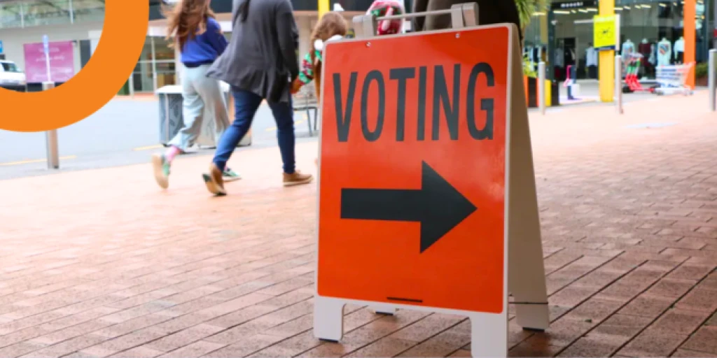 An orange voting sign on a busy footpath