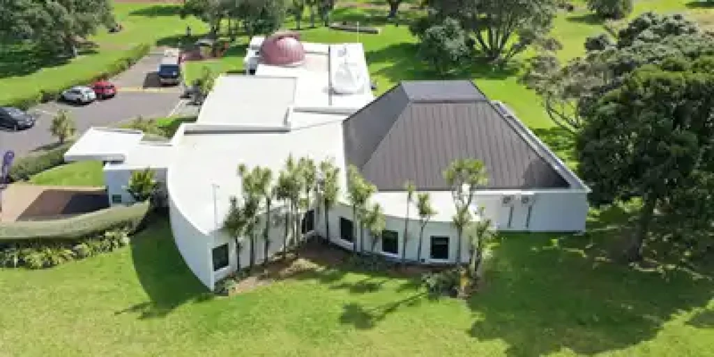 Aerial view of a white observatory with a triangular black roof and a dome, surrounded by greenery, trees, and a parking lot in a suburban park setting.