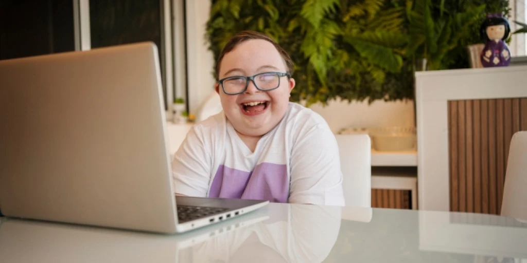 A boy with down syndrome sitting at a table, using a laptop