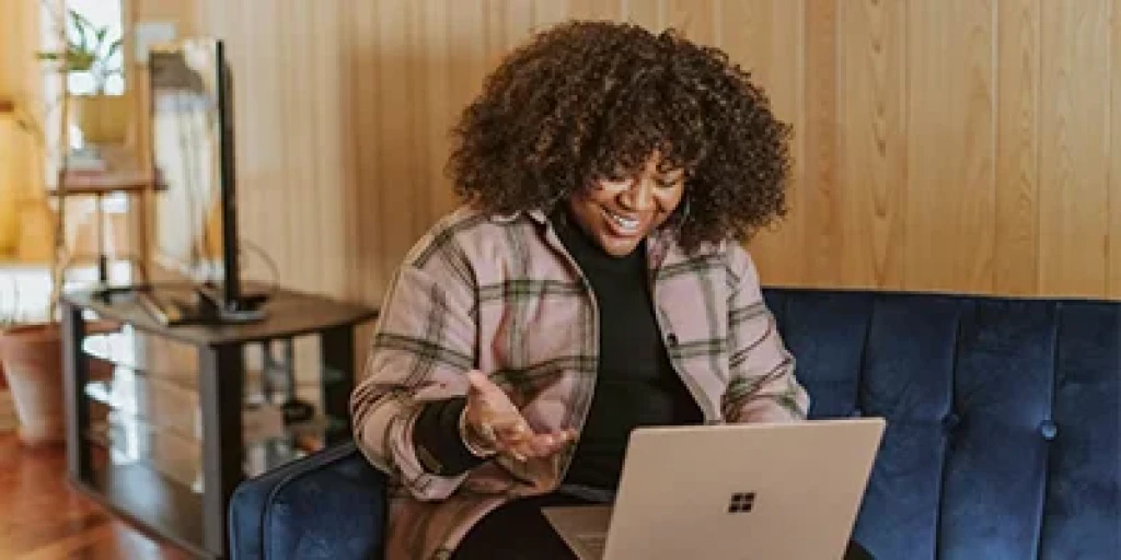 A smiling woman sitting on couch, looking at a laptop computer.