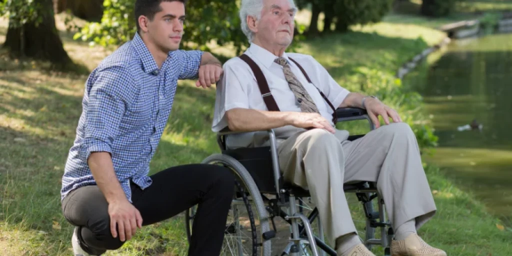 A young man crouching next to an elderly man in a wheelchair by a duck pond