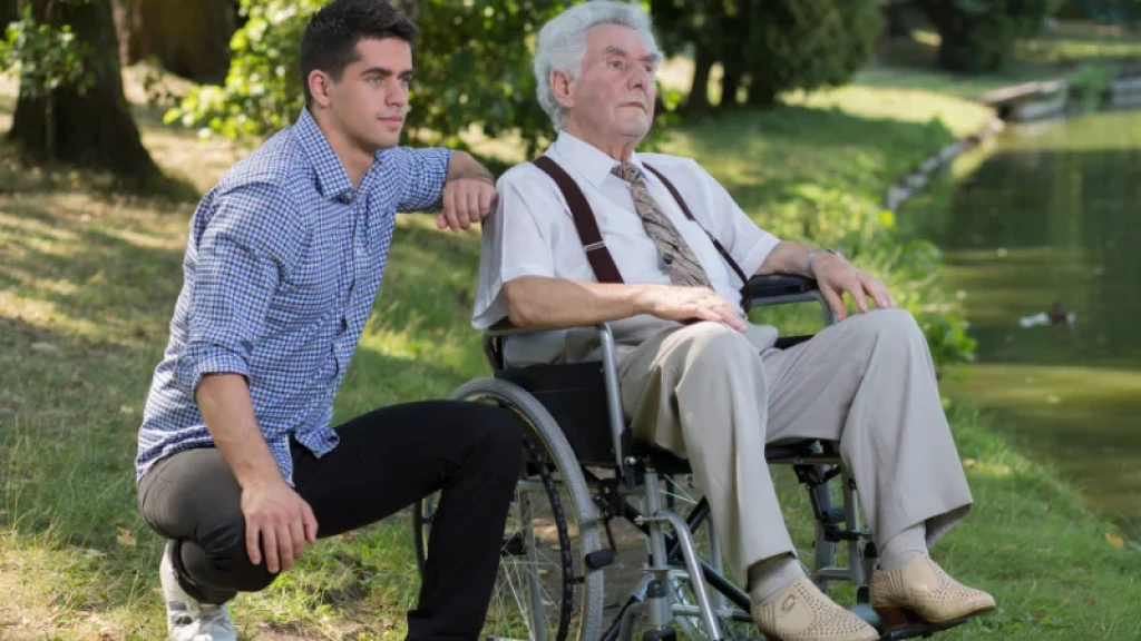 A young man crouching next to an elderly man in a wheelchair by a duck pond