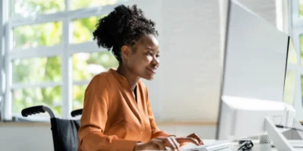 A woman wearing and orange shirt, sitting in a wheelchair, working on a computer