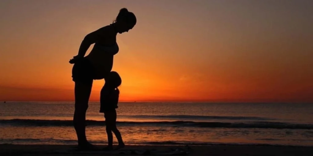 The silhouette of a small child kissing a pregnant woman's belly on a beach at sunset.