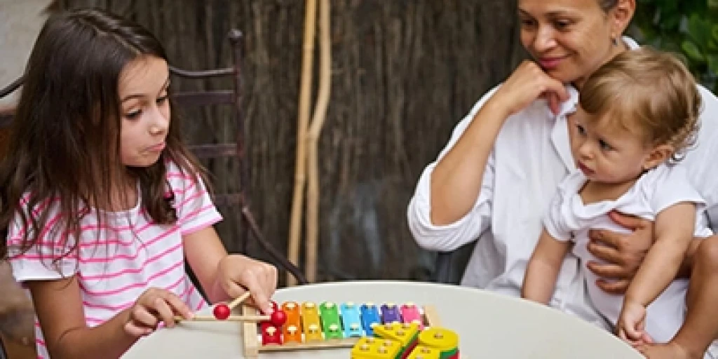 A girl plays with a rainbow xylophone while a woman and toddler watch.
