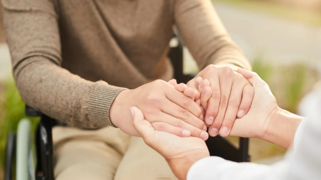 A wheelchair user in a brown sweater holding hands with a person in a white shirt