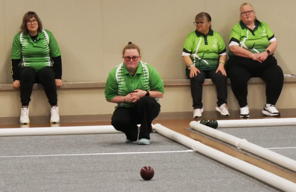 Lady in a green polo crouching, looking at the red bocce ball she just bowled